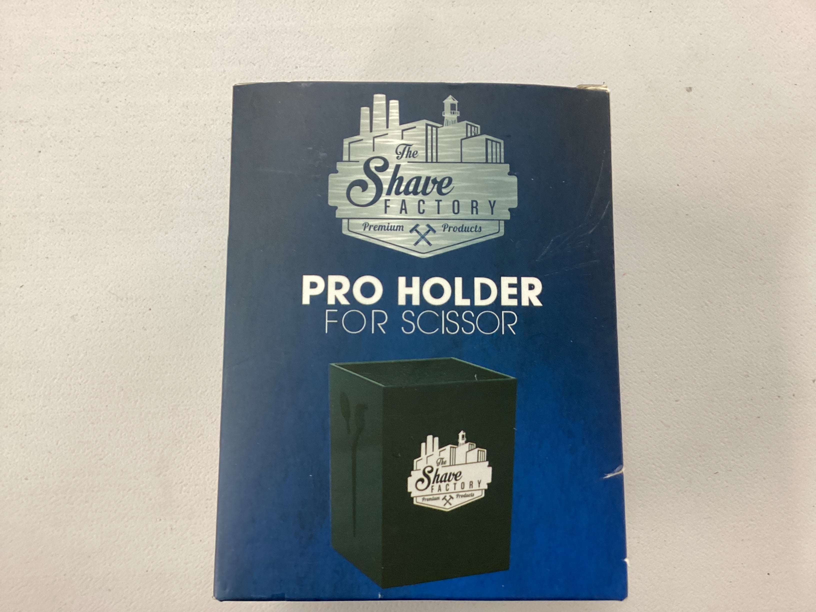 Blue packaging of 'The Shave Factory' pro holder for scissor on a beige background